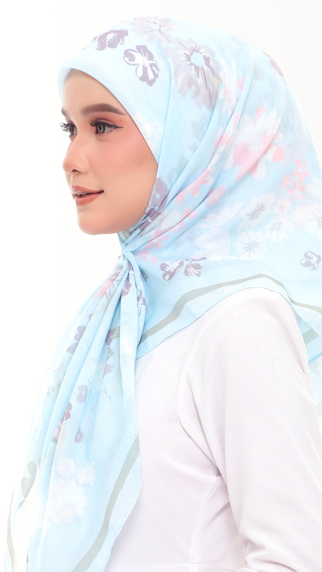 A woman wearing a light blue floral hijab with pink and gray accents, paired with a white long-sleeve top, shown in profile against a plain white background.