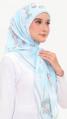 A woman wearing a light blue floral hijab with gray and pink accents, paired with a white long-sleeve top, set against a plain white background.