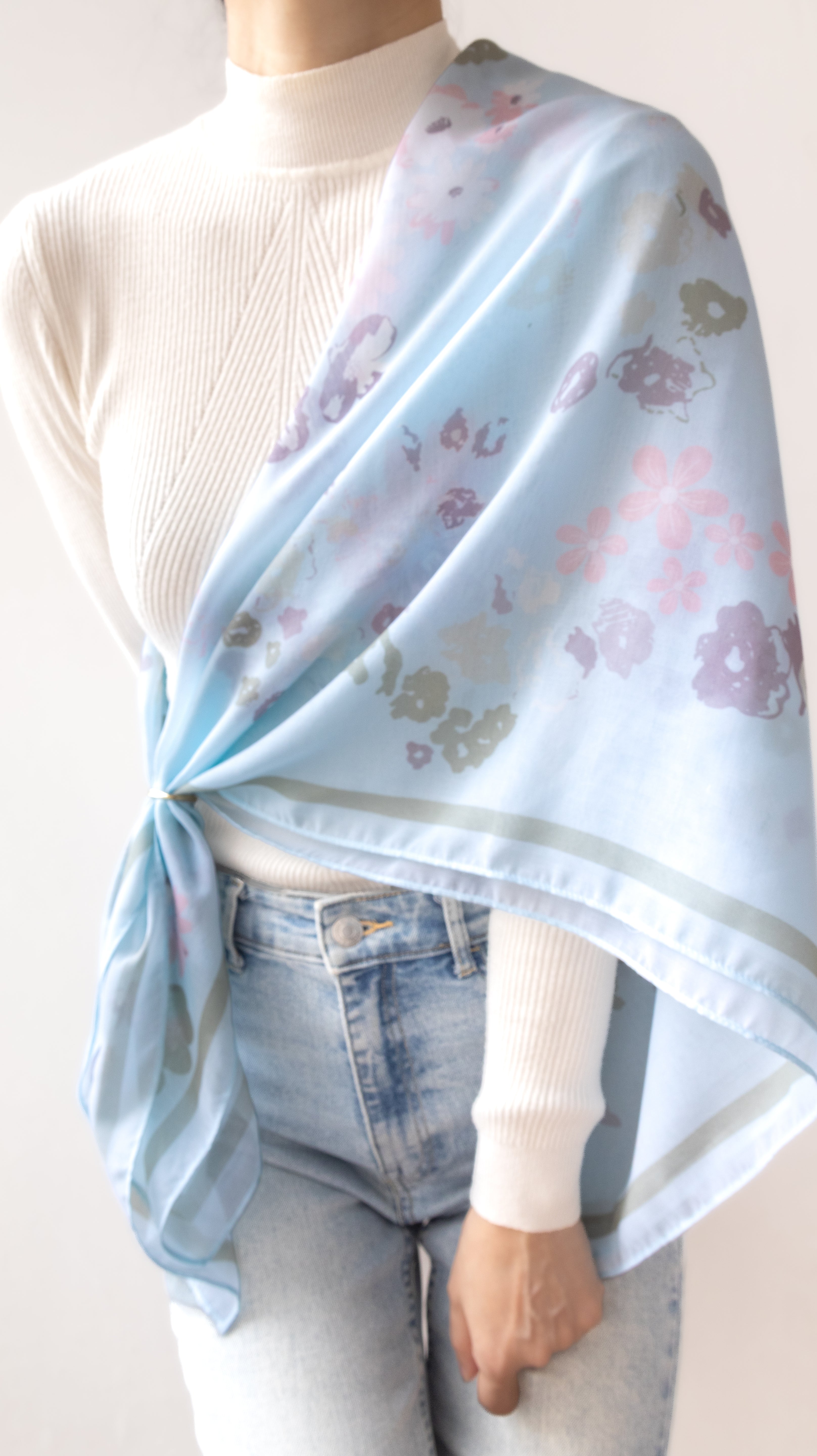 A light blue scarf with a delicate floral pattern in pink, purple, and green, styled over a white ribbed turtleneck sweater and light-wash jeans.