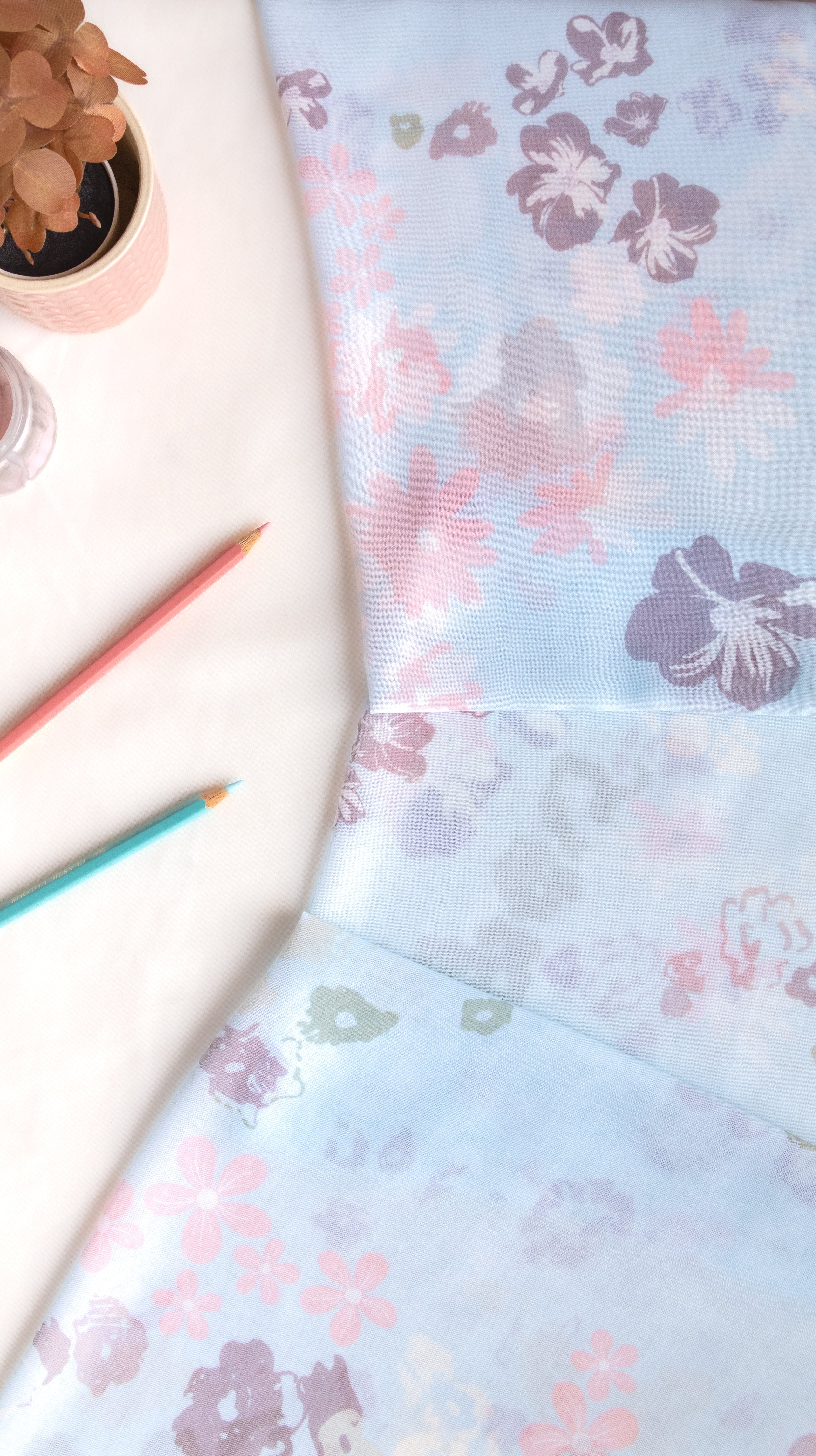 A folded piece of light blue fabric with a watercolor-style floral print in pink, purple, and green, styled on a white surface with a small potted plant, colored pencils, and a glass.