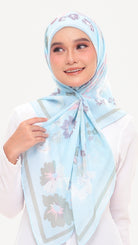 A woman wearing a light blue hijab with a floral print and gray border details, paired with a white long-sleeve top against a plain white background.