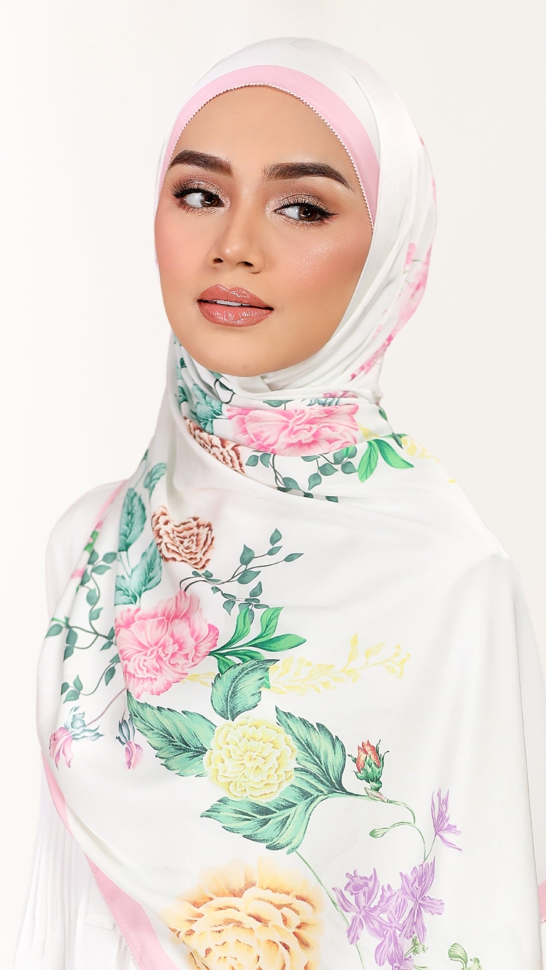 A woman wearing a white hijab with a pastel pink undercap and a colorful floral print featuring pink, yellow, and green roses and leaves, set against a plain white background.