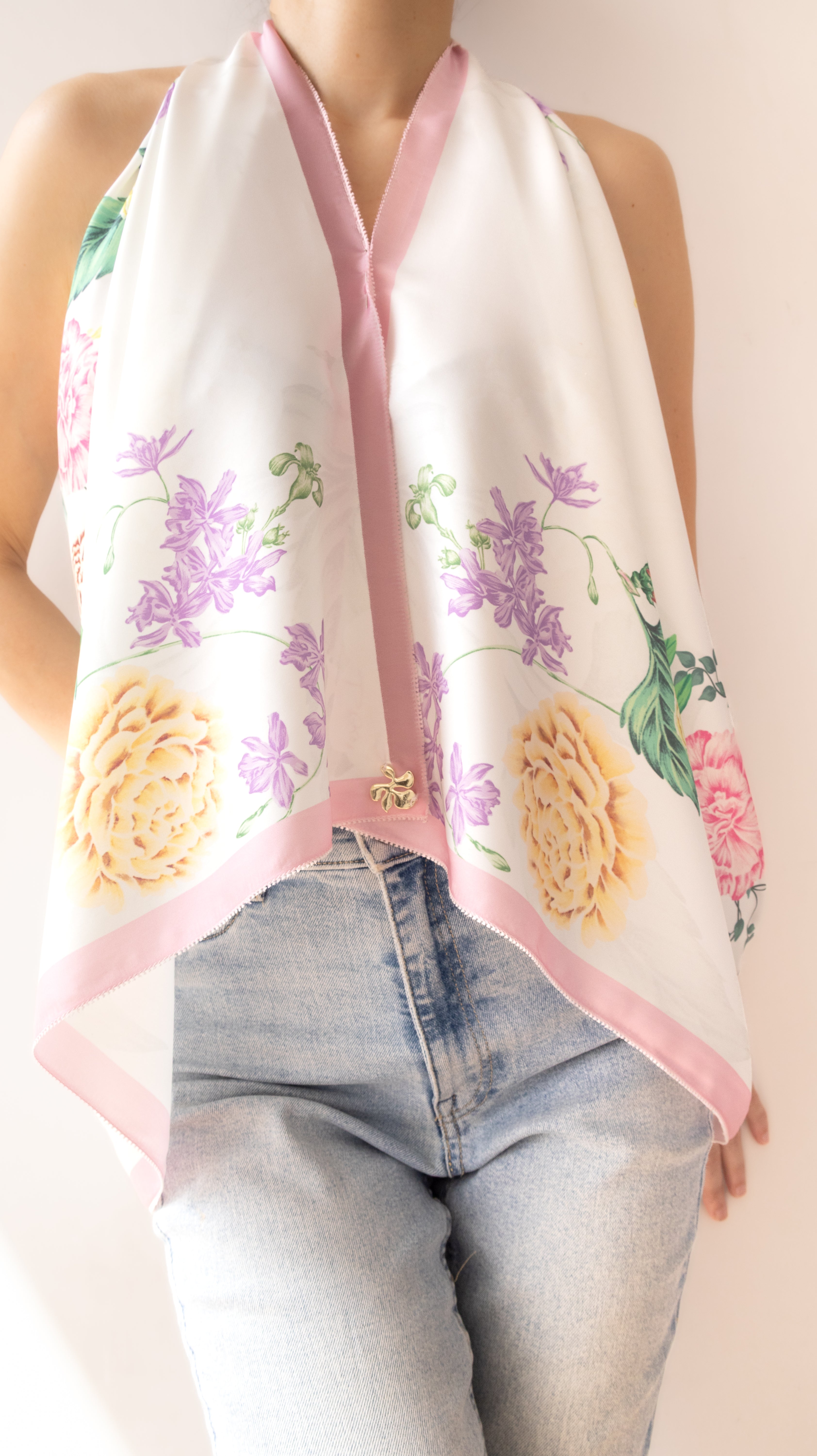 A white halter-style scarf with pink trim and a colorful floral print, worn by a model in light-wash blue jeans against a plain light background.