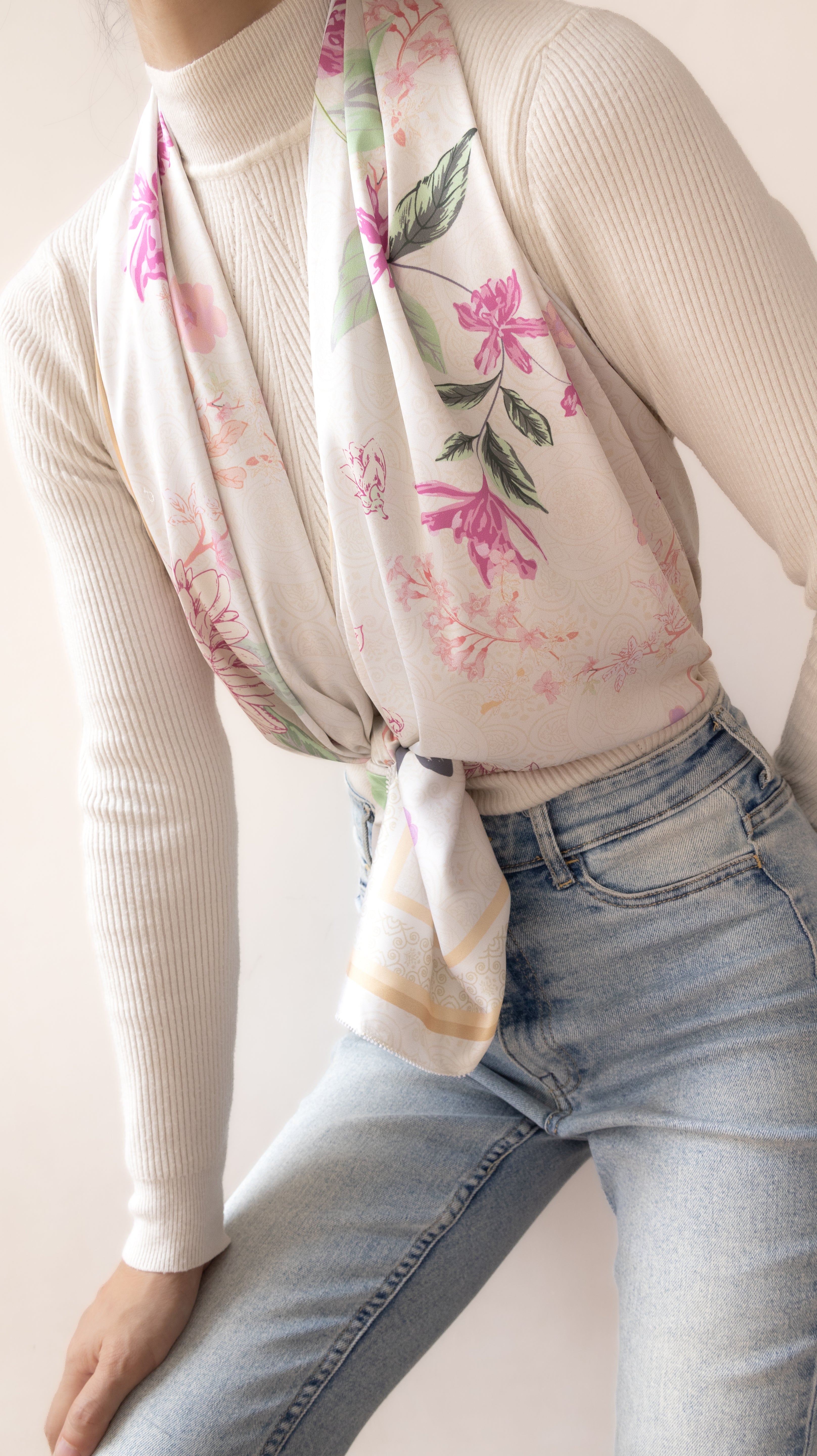 A cream-colored scarf with a pink and green floral print, styled tied around the waist of a model wearing a cream ribbed turtleneck sweater and light blue jeans.