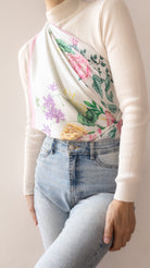 A person wearing a cream ribbed mock-neck top with a draped floral print panel over the chest, paired with light blue jeans, against a plain beige background.