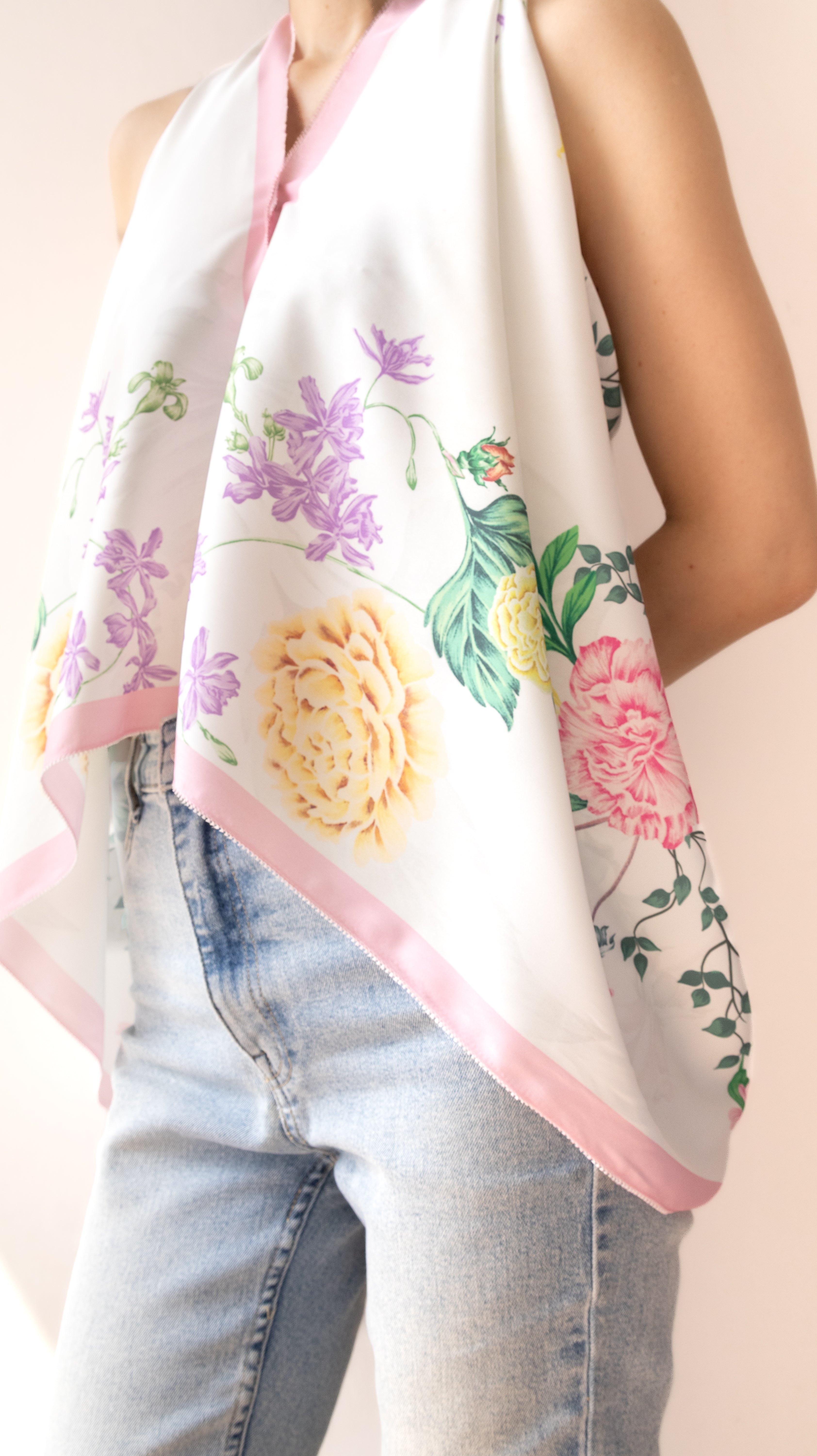 A white halter-style scarf with a pink border and a colorful floral print featuring yellow, pink, and purple flowers with green leaves, worn by a model in light-wash blue jeans.