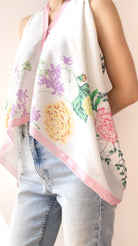A white halter-style scarf with a pink border and a colorful floral print featuring yellow, pink, and purple flowers with green leaves, worn by a model in light-wash blue jeans.