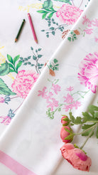 A folded white fabric with a pink and green floral print, styled with two colored pencils and pink flowers on a white background.