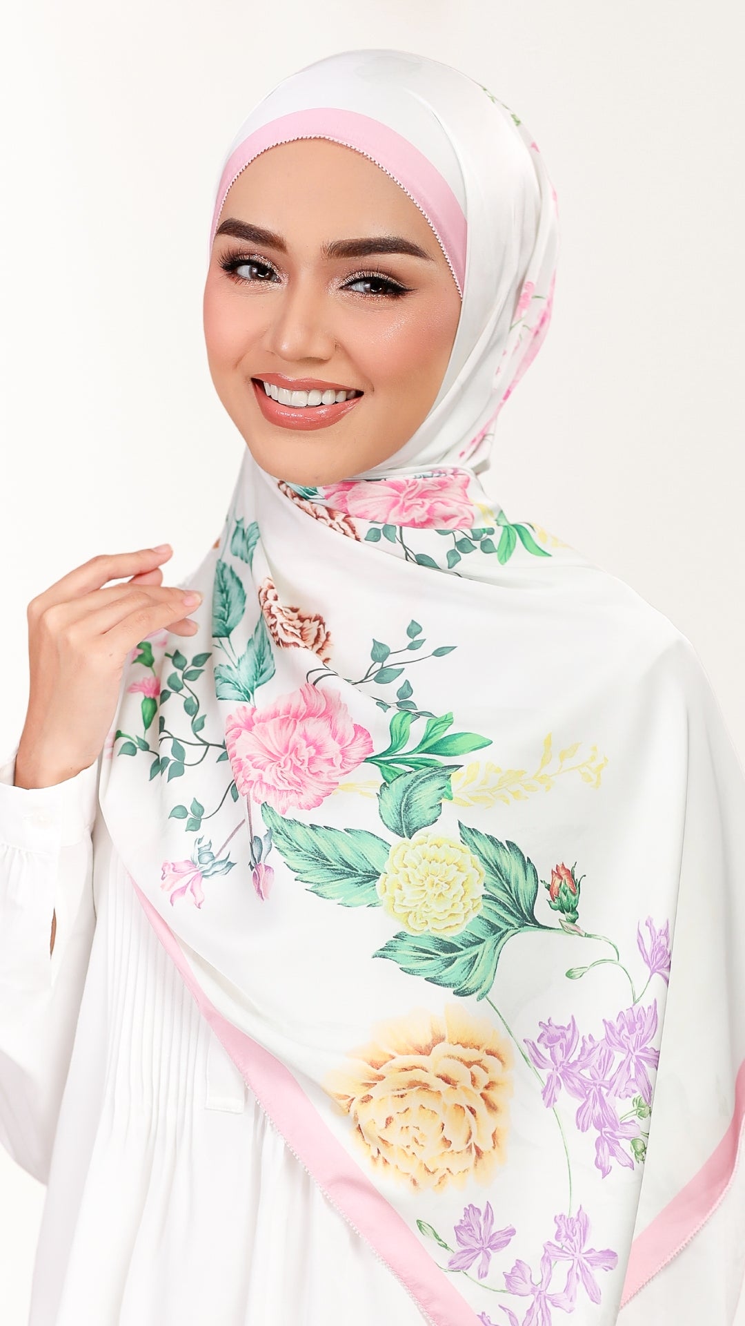 A woman wearing a white hijab with a pink band and a colorful floral print featuring pink, yellow, and purple flowers with green leaves, set against a plain white background.