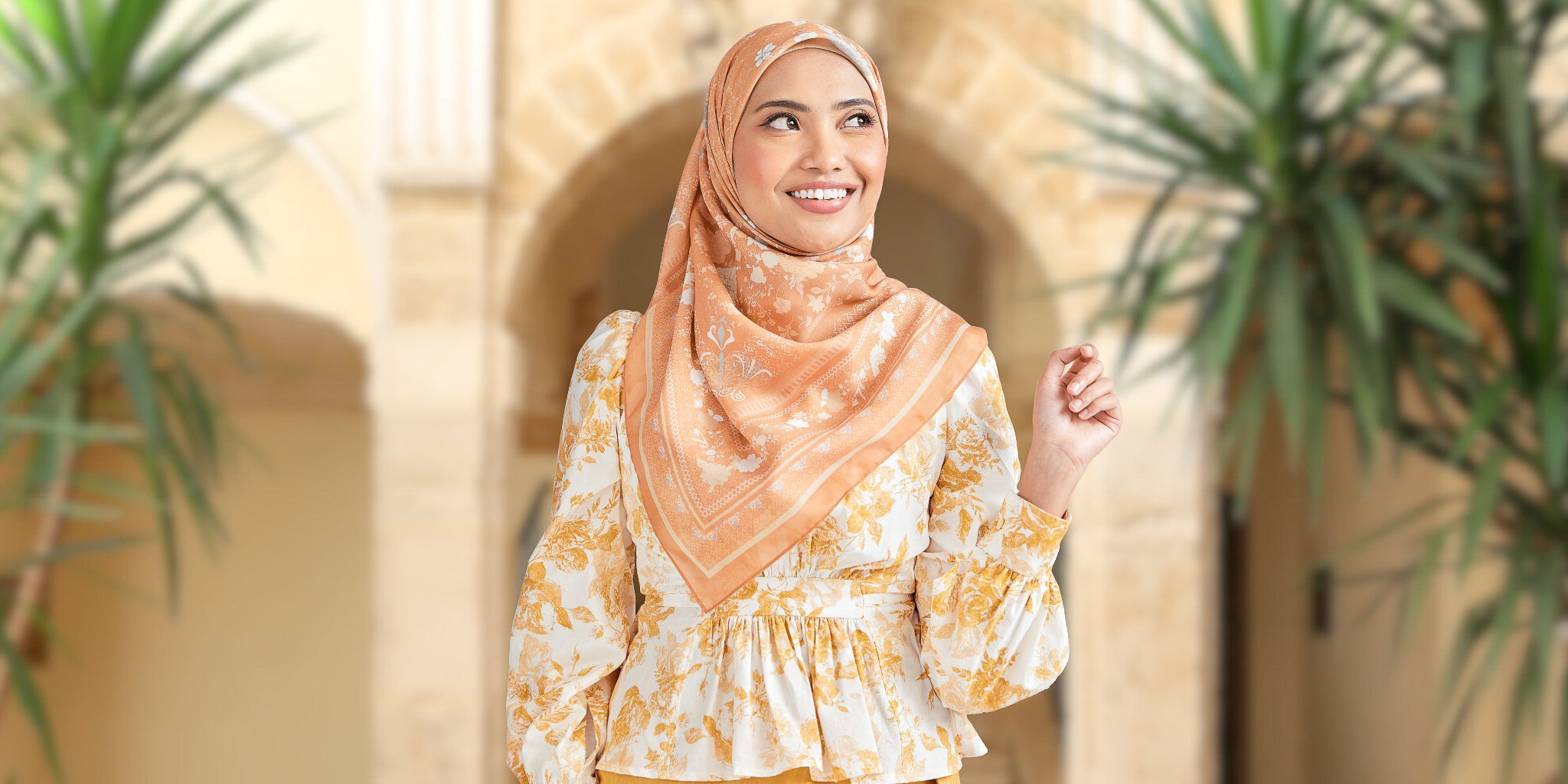 Woman wearing a floral dress and hijab standing outdoors with plants and a building in the background