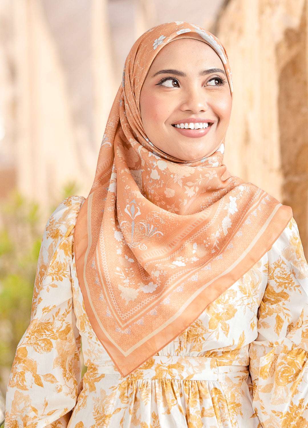 Woman wearing a peach hijab with floral patterns against a blurred natural background