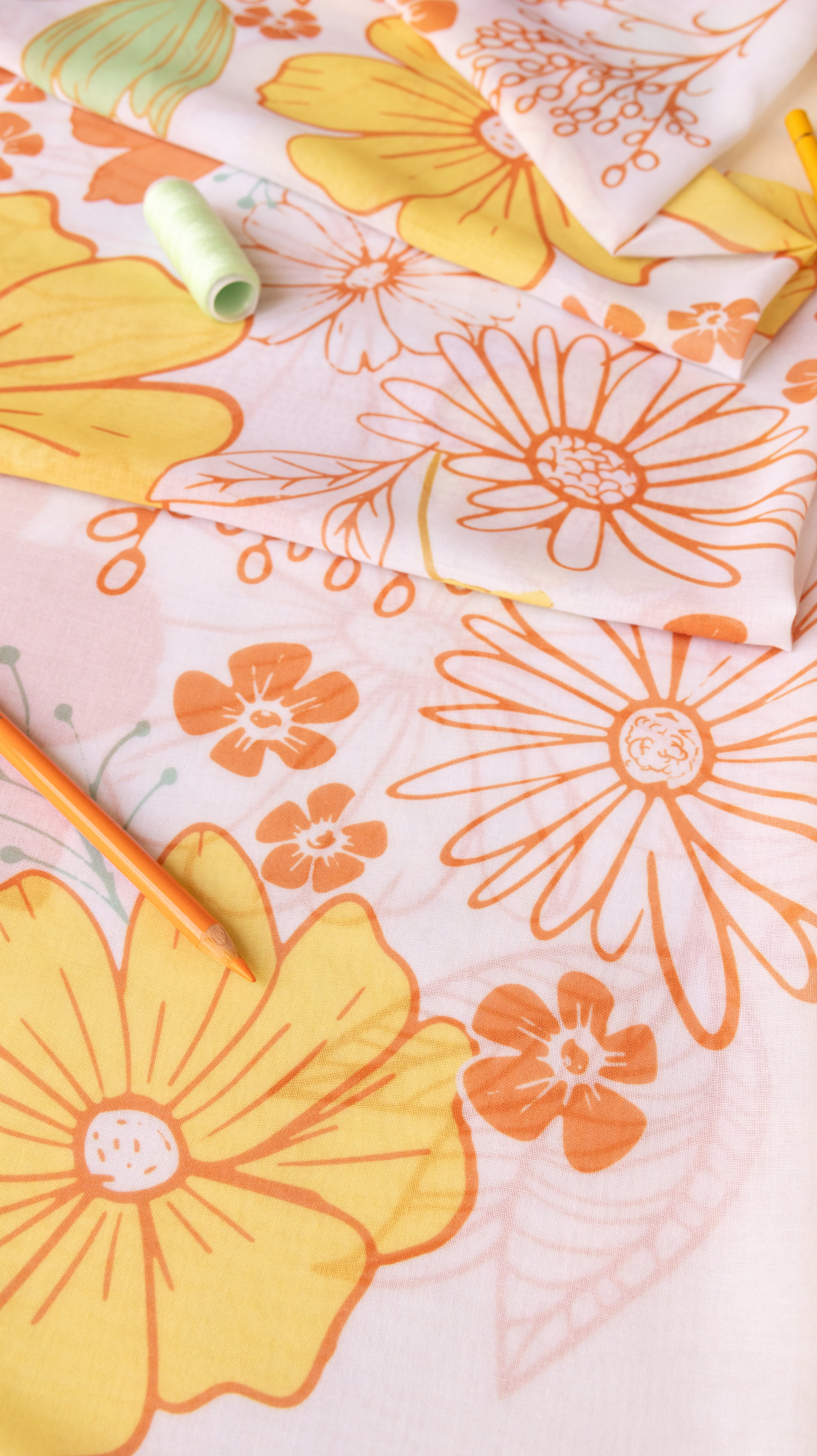 A close-up of a light pink fabric with a retro floral pattern in orange and yellow, styled with a spool of light green thread and orange pencils.