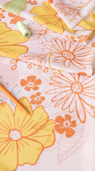 A close-up of a light pink fabric with a retro floral pattern in orange and yellow, styled with a spool of light green thread and orange pencils.