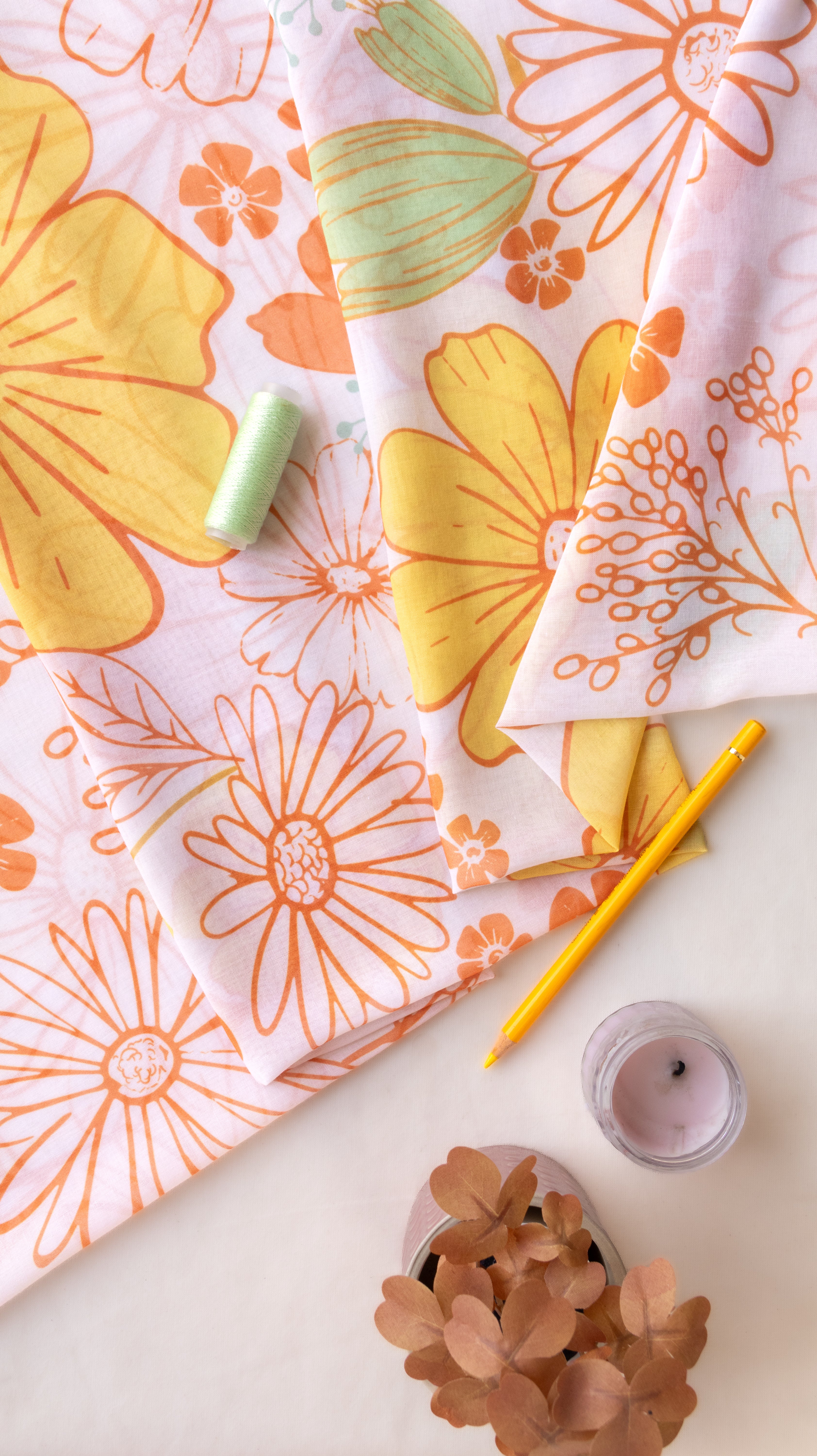 A styled flat-lay of two patterned fabrics with retro daisy and floral designs in orange, yellow, and mint green on a white background, accompanied by a pencil, thread, candle, and dried flowers.