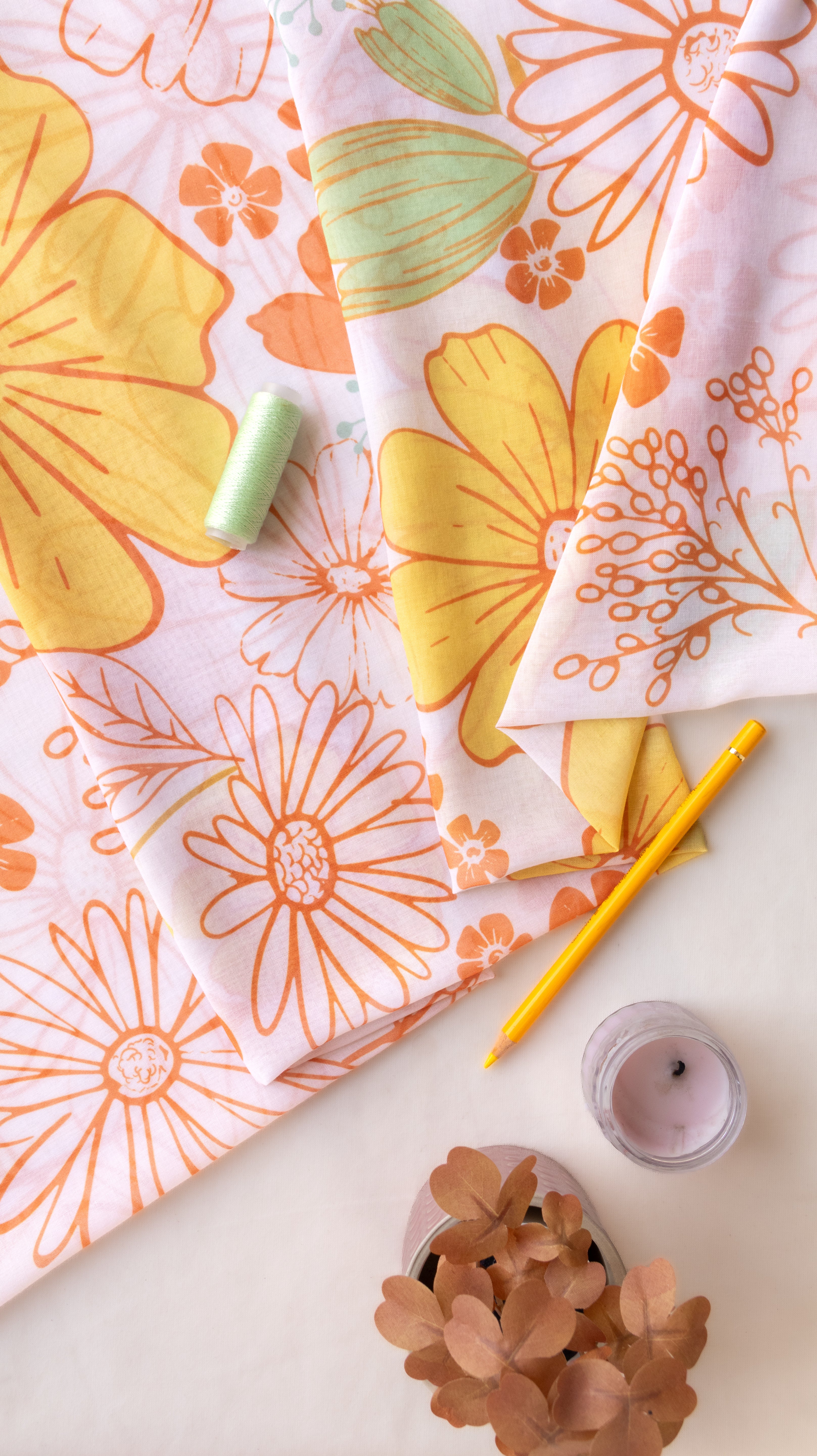 A styled flat-lay of two patterned fabrics with retro daisy and floral designs in orange, yellow, and mint green on a white background, accompanied by a pencil, thread, candle, and dried flowers.