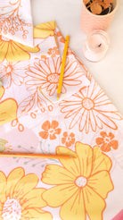 A styled flat-lay of a floral fabric or paper with large yellow and orange daisy-like flowers on a white background, alongside two yellow pencils and small decorative containers.