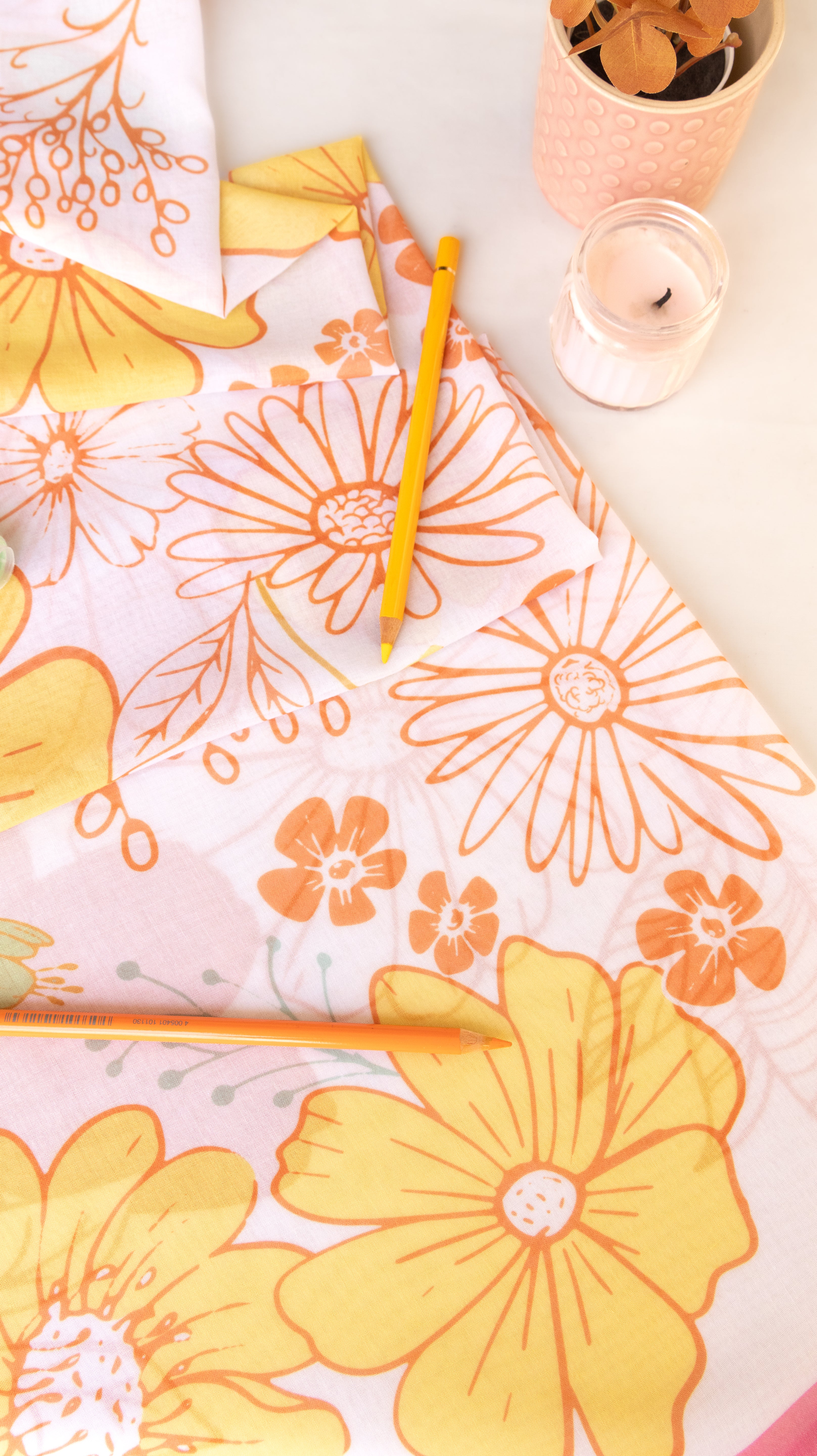 A styled flat-lay of a floral fabric or paper with large yellow and orange daisy-like flowers on a white background, alongside two yellow pencils and small decorative containers.