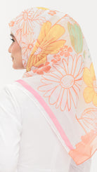 A woman shown from the side wearing a sheer white hijab with a colorful floral print in yellow, orange, and peach, featuring a pink border.