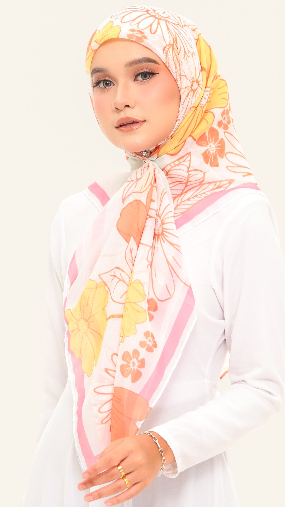 A woman wearing a white long-sleeve top and a light pink hijab with a yellow and orange floral pattern, posed against a solid off-white background.