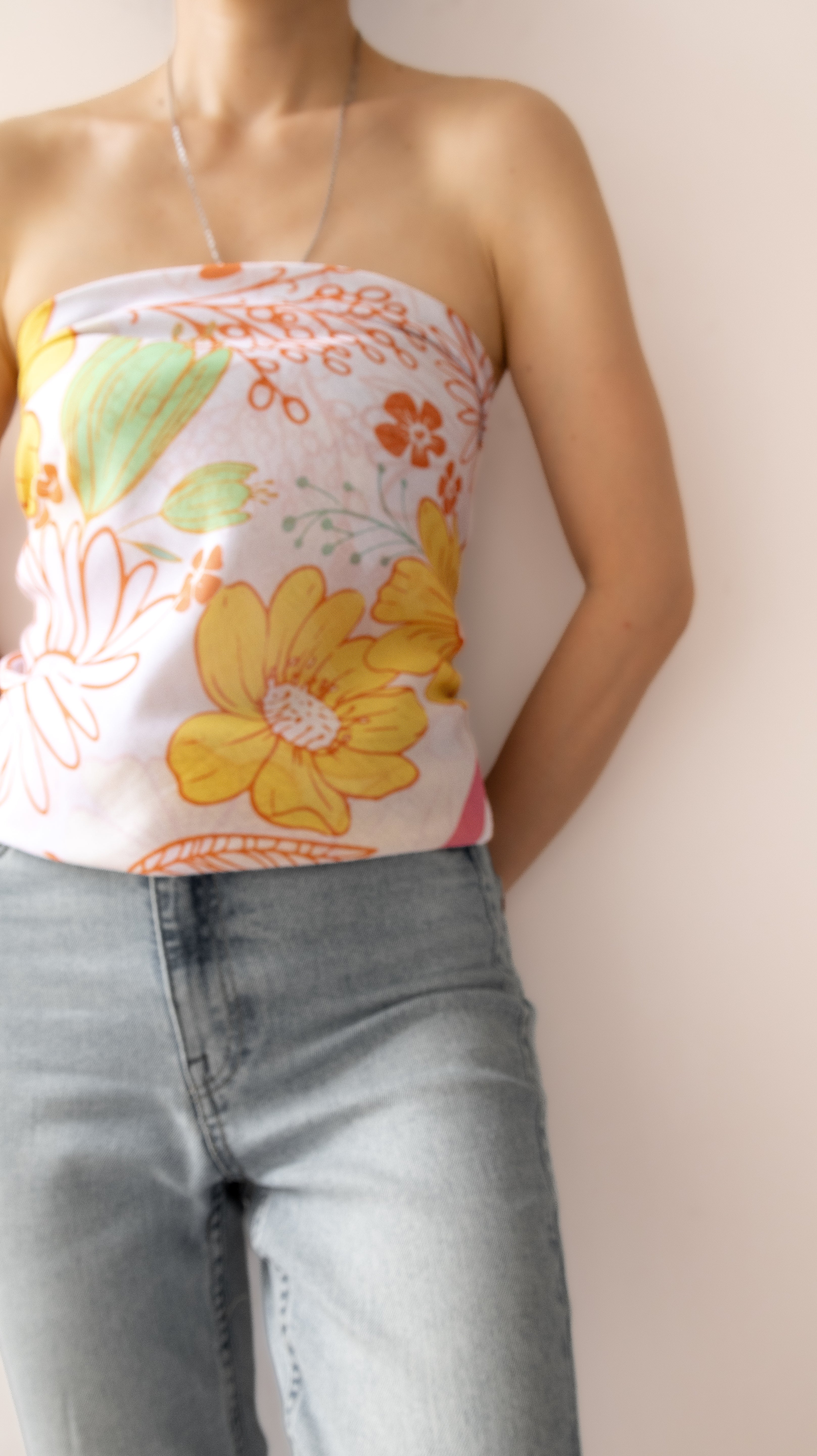A woman wearing a strapless tube top with a retro floral print in yellow, orange, and green on a white background, paired with light-wash blue jeans.