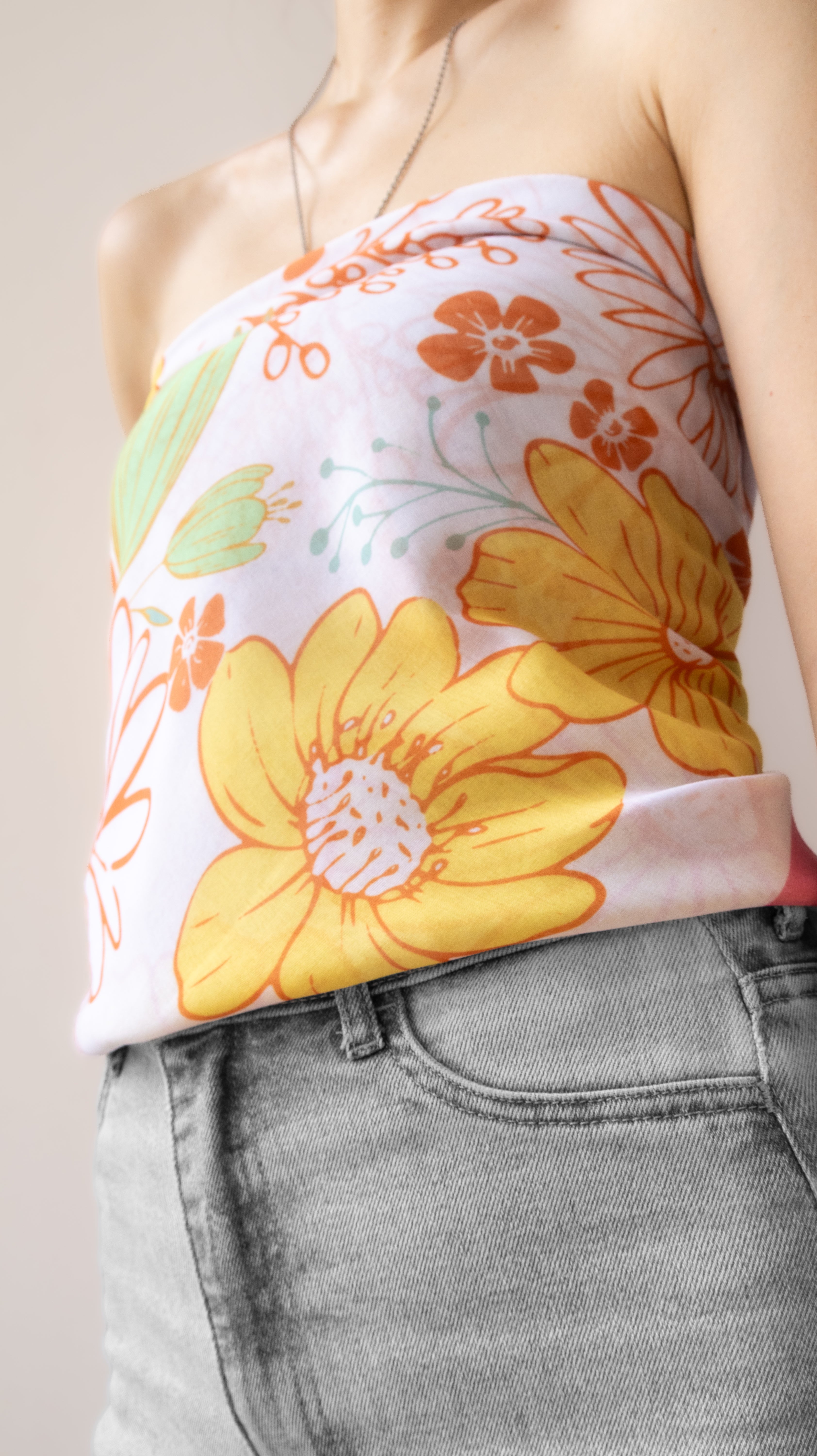 A close-up of a woman wearing a strapless tube top with a retro floral print in yellow, orange, and green on a white base, paired with light gray denim jeans.