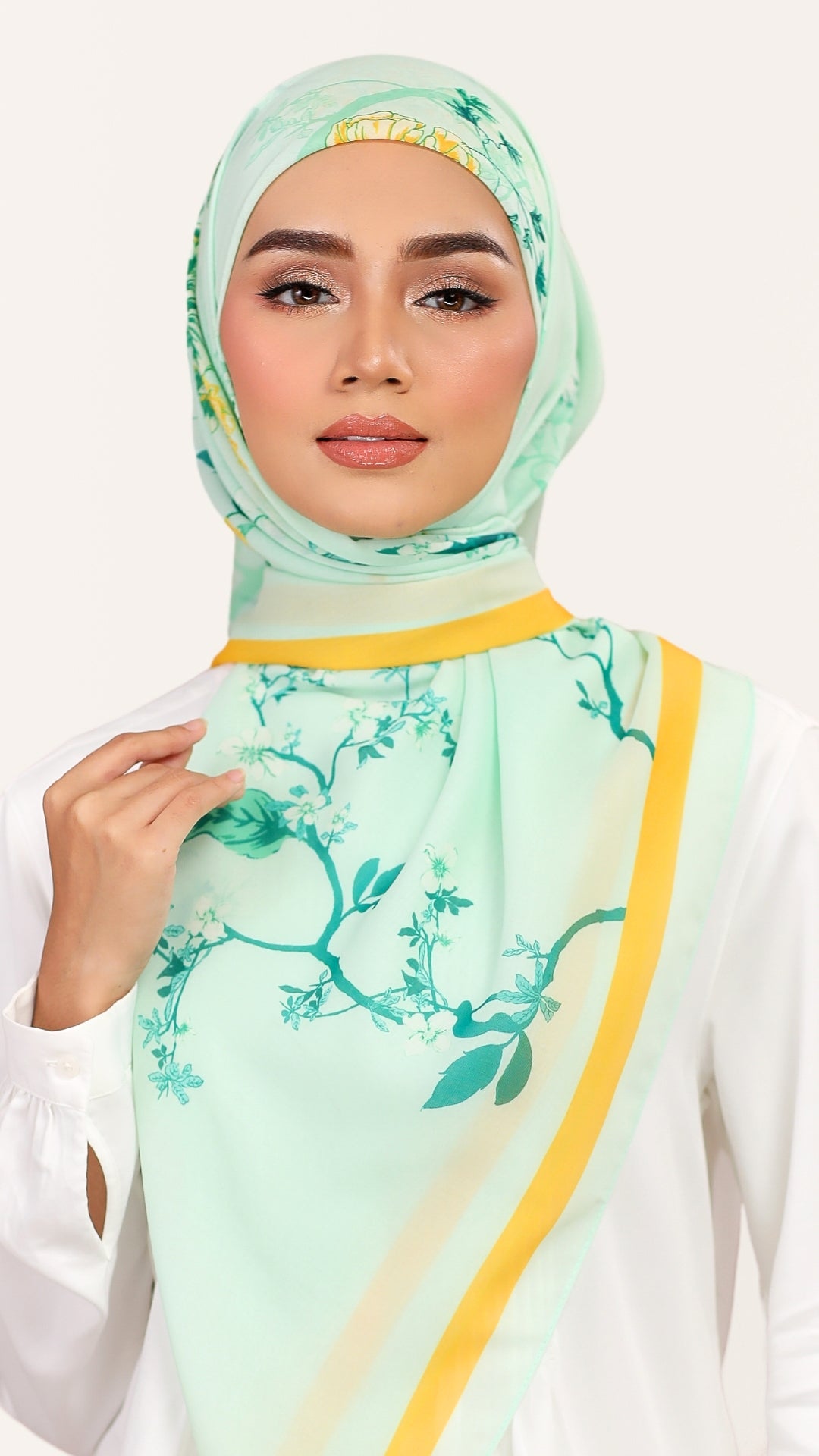 A woman wearing a mint green hijab with a teal floral vine print and yellow stripe accents, paired with a white top against a light off-white background.