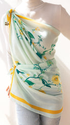 A woman wearing a white ribbed long-sleeve top and light blue jeans, wrapped in a mint green silk scarf with yellow floral patterns and yellow trim.