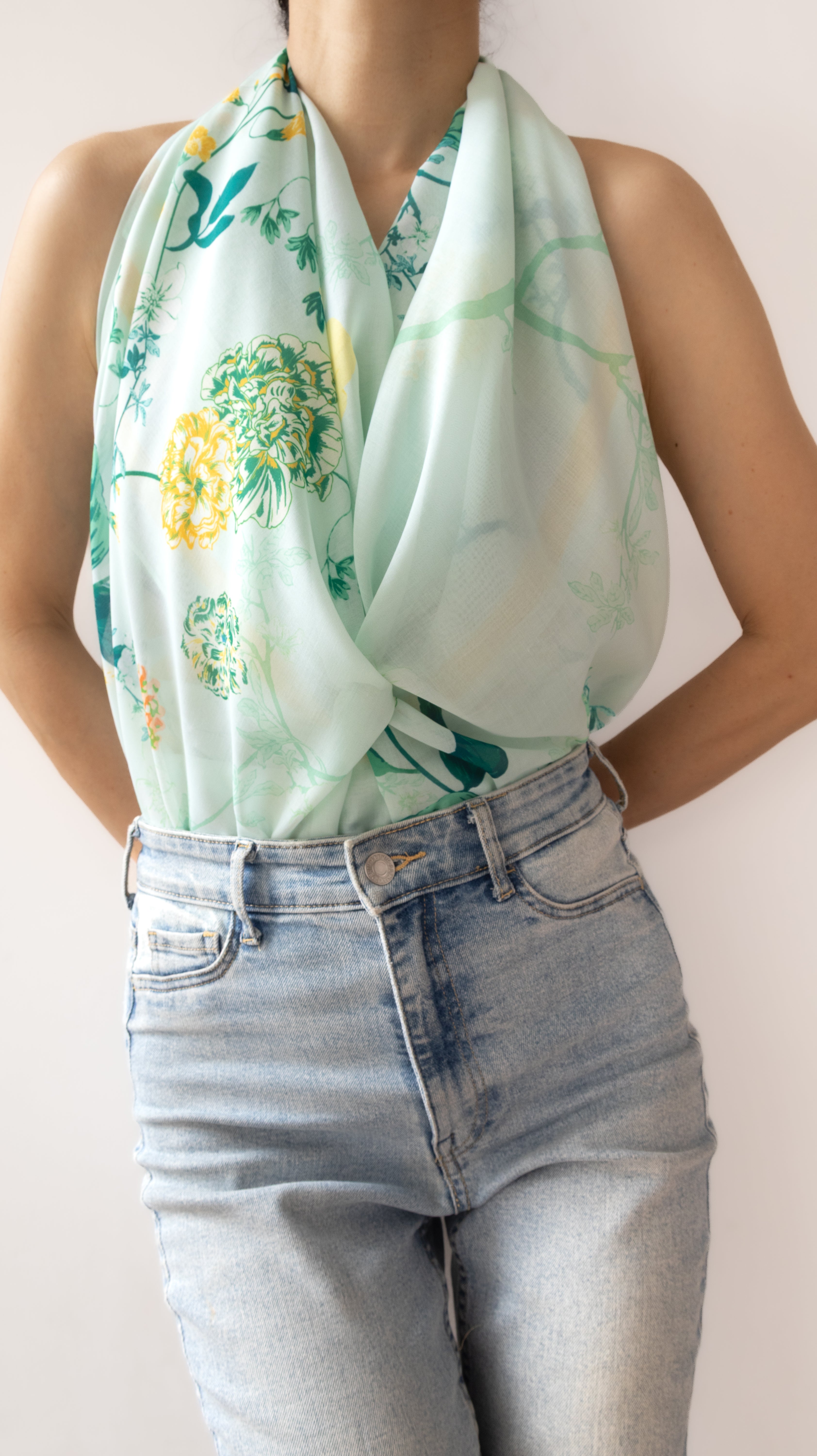 A person wearing a light mint green halter-style scarf with a yellow and green floral print, paired with light-wash blue jeans against a plain light background.
