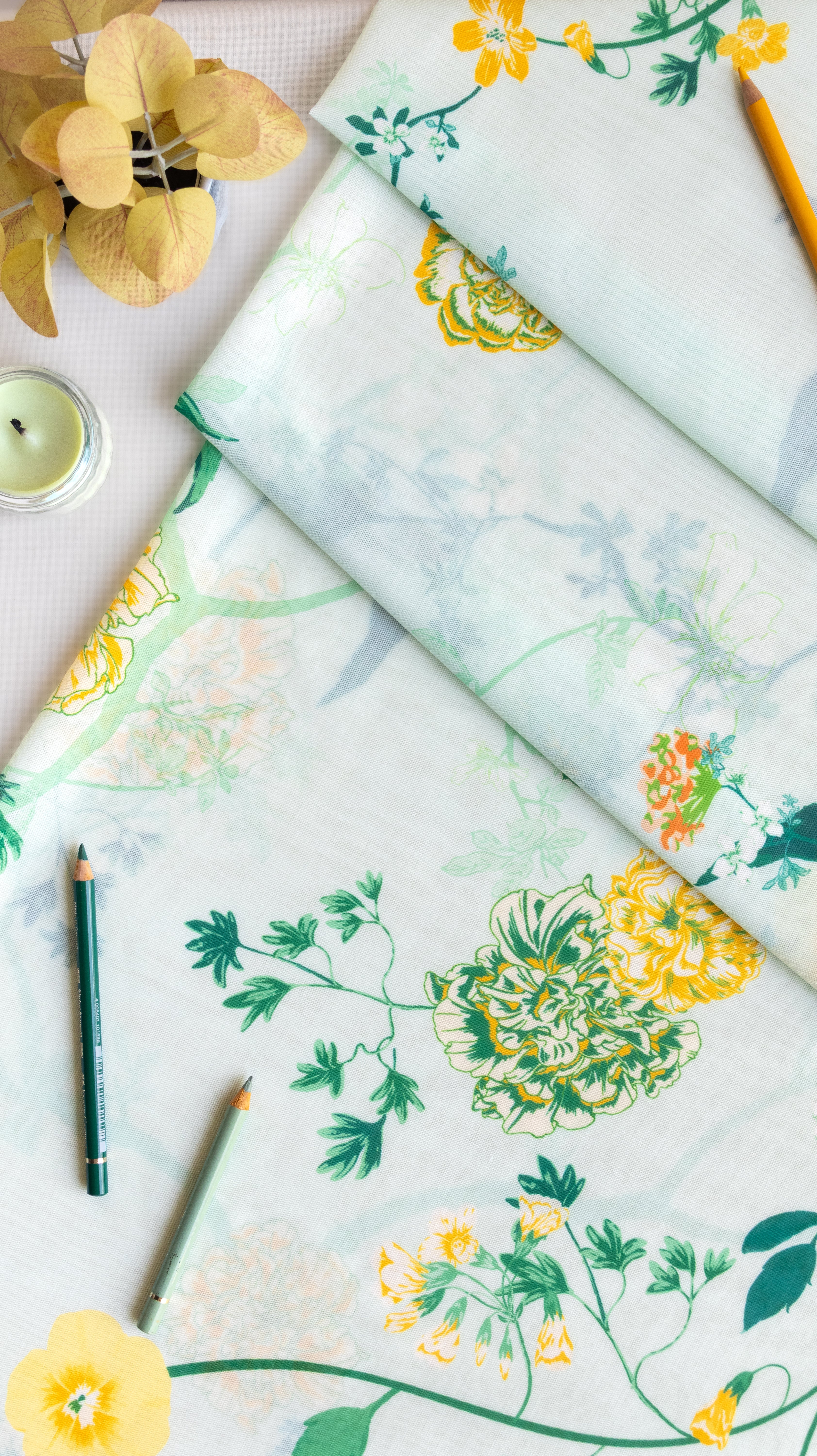 A folded and flat-laid piece of fabric with a botanical floral print, styled with pencils, a candle, and dried flowers on a white background.