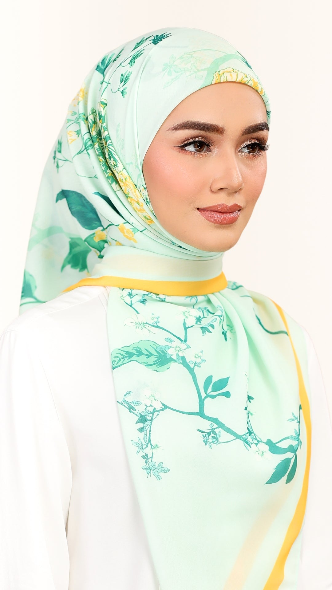 A woman wearing a mint green hijab with a delicate floral branch print and yellow-gold trim, paired with a white top against a clean white background.