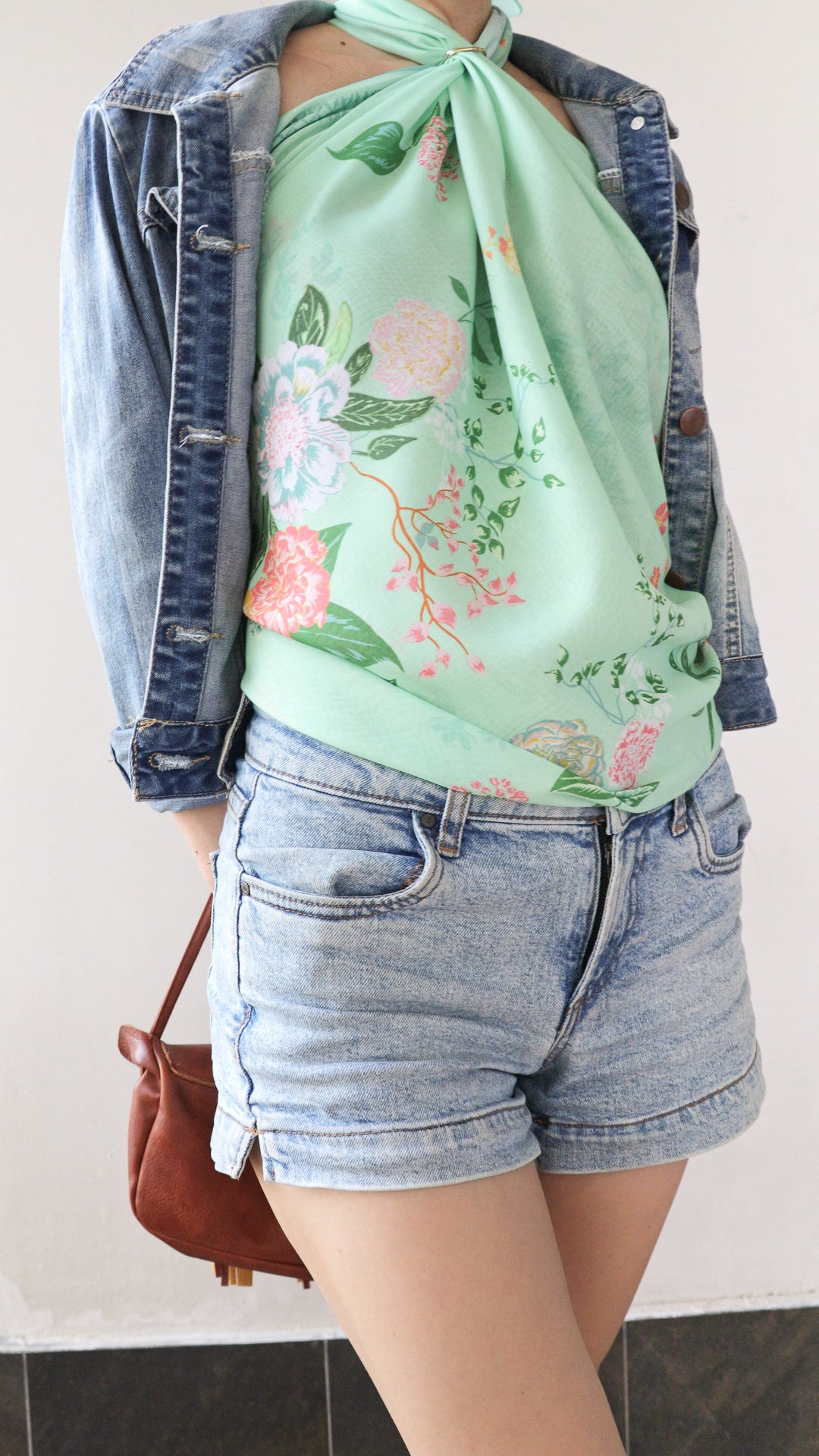 A person wearing a mint green floral halter top with a denim jacket and light-wash denim shorts, holding a brown handbag.