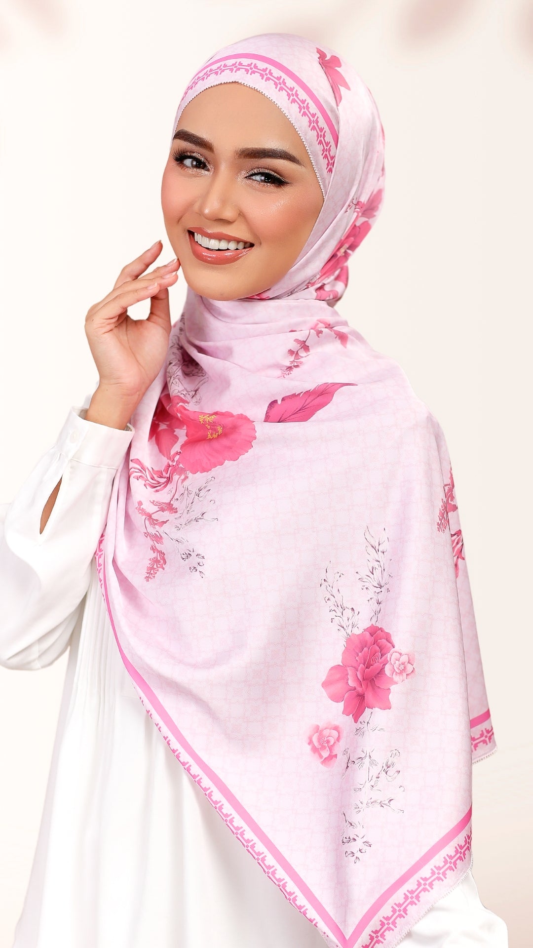 A smiling woman wearing a light pink hijab with a darker pink floral print and delicate border trim, paired with a white long-sleeve top against a soft off-white background.