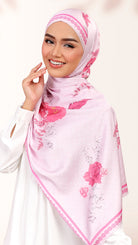 A smiling woman wearing a light pink hijab with a darker pink floral print and delicate border trim, paired with a white long-sleeve top against a soft off-white background.