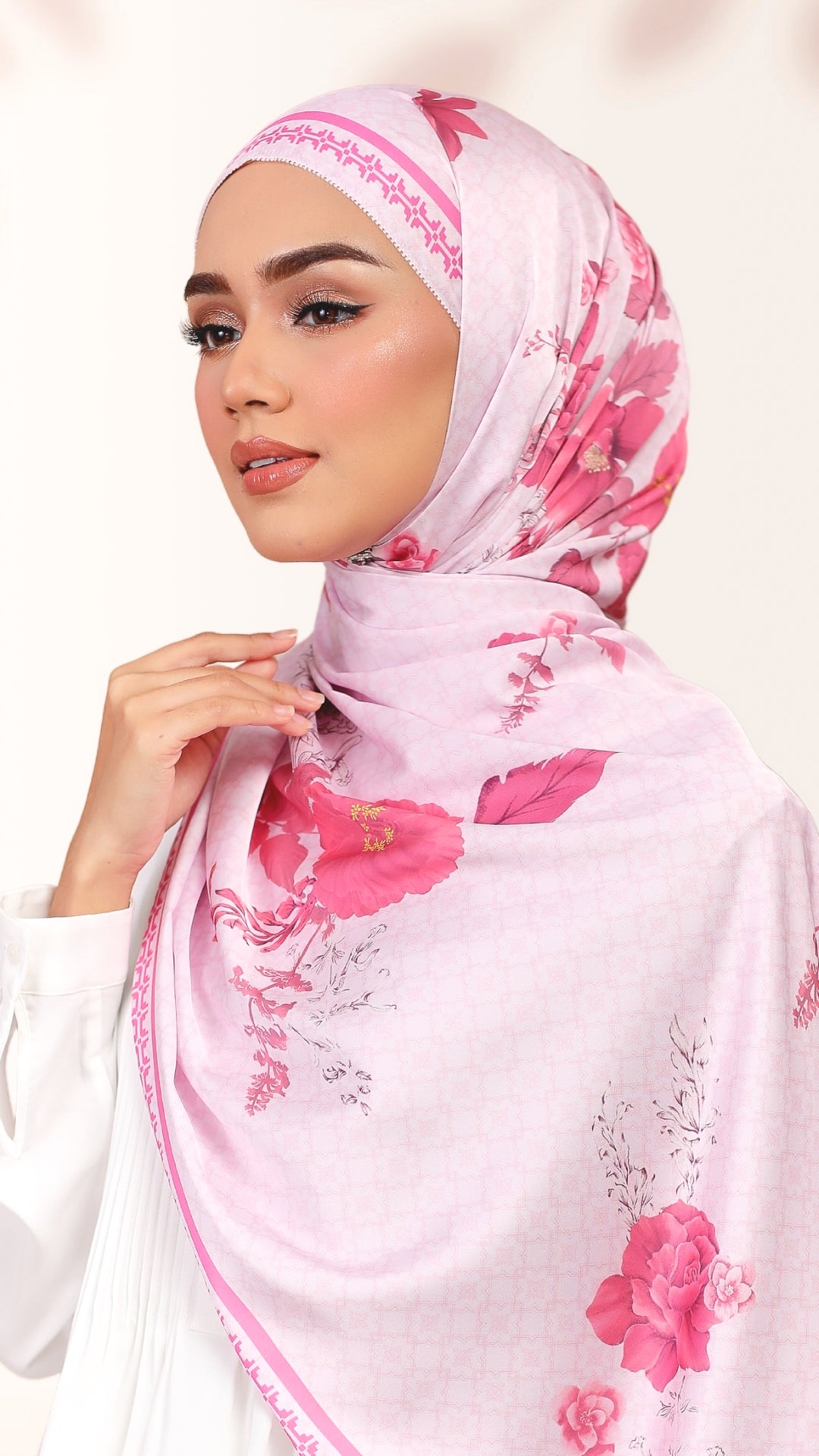 A woman wearing a light pink hijab with a vibrant pink floral print and delicate embroidered trim along the edges, set against a soft, light beige background.