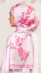 A woman wearing a light pink hijab with a vibrant pink floral print and decorative borders, shown in profile against a soft beige background.